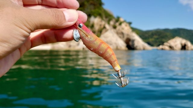 磯の海を背景に、指でつまんだオレンジのエギ