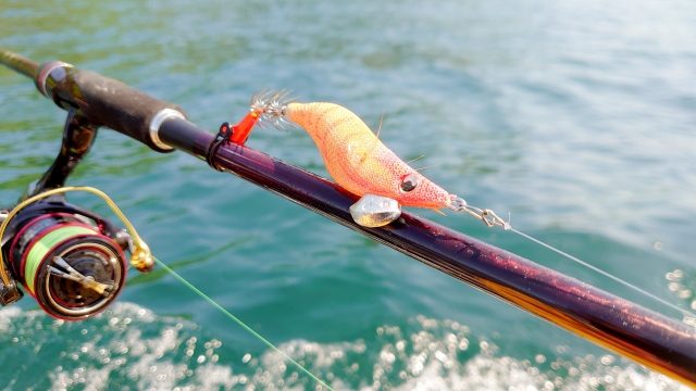 青い海を背景に、エギングロッドに掛けたオレンジのエギとスピニングリール