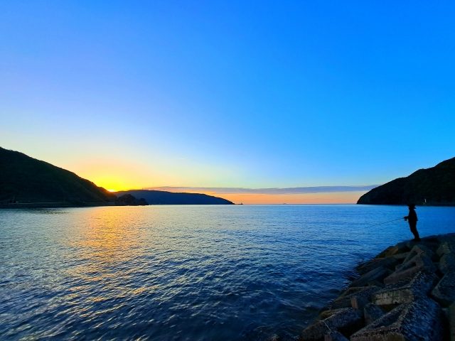 朝の湾と磯場に立つ釣り人のシルエット