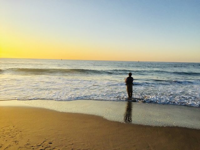 夕暮れのサーフで波打ち際に立ち、ルアーを投げる釣り人