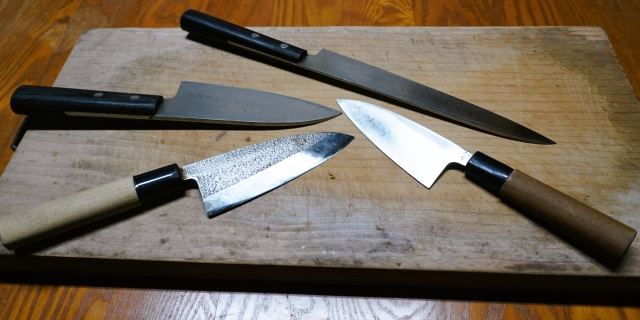 木のまな板に並べた4本の包丁。奥に細長い柳刃包丁と黒い柄の洋包丁、手前に木の柄の出刃包丁が2本