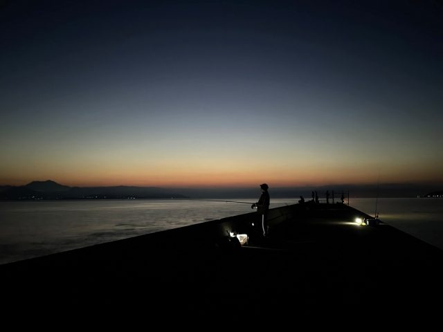 朝焼けの空の下、人のいない堤防の先端で竿を構える釣り人のシルエット。足元に常夜灯の明かりが2つ灯っている